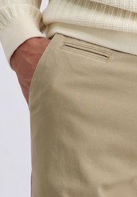 Beige, strukturierte Hose mit einer eleganten Seitentasche. Ein cremefarbener, gerippter Pullover ist sichtbar, mit einem eng anliegenden Bündchen. Lässige Stoffmischung.