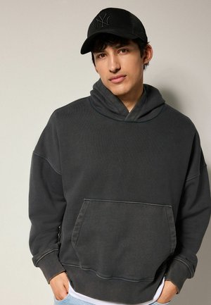 Dark gray hoodie with a large front pocket, funnel neck, and ribbed cuffs. Paired with a black cap featuring a logo in front.