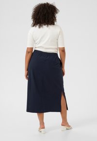 Fustă maxi bleumarin cu fanta laterală, talie elastică și material ușor. Modelul poartă un top alb cu mânecă scurtă și sandale cu spatele deschis.
