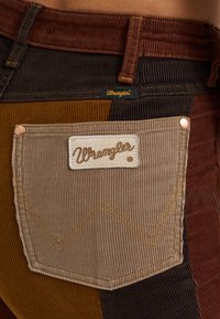 Brown corduroy jeans featuring a multi-colored patchwork design, finished with a beige pocket, Wrangler logo, and copper rivets.