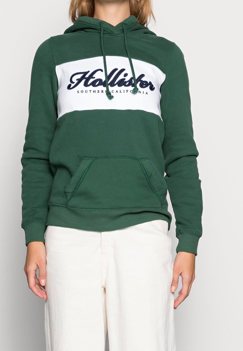 Zelená mikina s kapucí vyrobená z bavlněné směsi s bílým vodorovným pruhem. Obsahuje logo "Hollister", přední klokaní kapsu a šňůrku na kapuci.