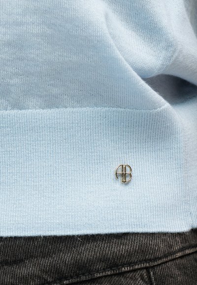 Hellblauer Strickstoff mit einer kleinen metallischen Logo-Brosche nahe dem Saum, oberhalb eines dunklen, strukturierten Stoffes.