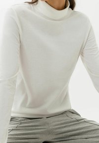 Person, die ein weißes langärmeliges Turtleneck-Shirt und graue strukturierte Hose trägt, sitzt vor einem schlichten weißen Hintergrund.