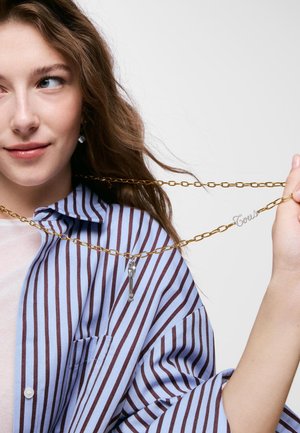 Joven mujer con cabello castaño ondulado, vistiendo una camisa a rayas azul y marrón, sosteniendo un collar de cadena dorada con un colgante de llave y un charm de "Tous".
