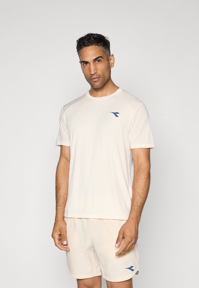 Man standing against plain background wearing light beige sportswear with blue logos on chest and shorts.