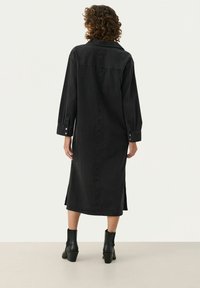 Czarna dżinsowa sukienka z długimi rękawami, guzikami przy mankietach, bocznymi rozcięciami i luźnym krojem, sięgająca do połowy łydki. Noszona z botkami do kostek.