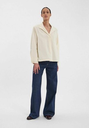 Chemise beige oversize avec un col en V, associée à un jean large bleu foncé. Tissu doux avec une coupe décontractée, sans motifs visibles.