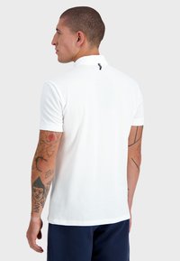 Chemise polo blanche avec un col rabattu et un petit accent au dos. Manches courtes et texture lisse. Tatouages visibles sur les bras.