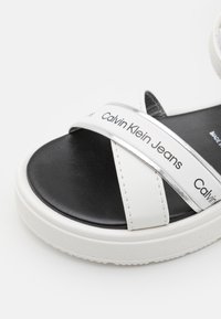Vita och svarta sandaler med korsade remmar, prydda med ett glänsande logoband, texturerad sula och platt design.