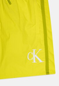 Shorts jaunes vifs en matériau léger, avec une bande latérale verte et un logo "ck" brodé en blanc à l'avant.