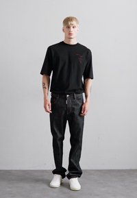 Un homme blond se tient de face, portant un t-shirt noir surdimensionné, un jean sombre, des baskets blanches et des tatouages visibles sur les deux avant-bras.