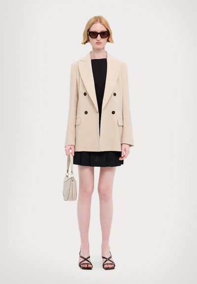 Blazer beige à double boutonnage, robe noire, jupe plissée noire, sandales noires à talons, et sac à main clair. Modèle portant des lunettes de soleil oversize.