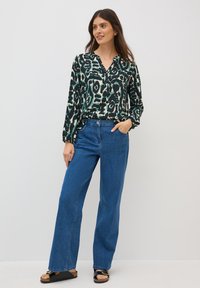 Blouse à motifs verts et crème avec un col en V, manches longues, associée à un jean large bleu et des sandales noires avec des semelles en liège.