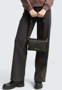 Schwarze Glitzer-Crossbody-Tasche mit rechteckiger Form, glänzender Textur und verstellbarem Schulterriemen, gehalten von einer Person, die dunkle Jeans und schwarze Schuhe trägt.