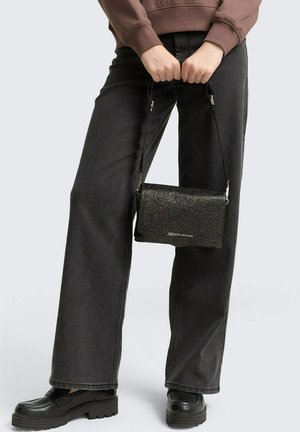 Black glitter crossbody bag with a rectangular shape, shiny texture, and adjustable strap, held by a person wearing dark jeans and black shoes.