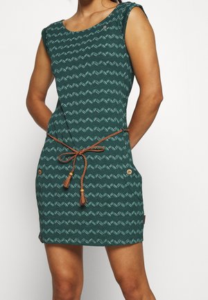 Mouwloze groene jurk met blauw geometrisch patroon, bruine gevlochten riem gestrikt in de taille en knoopdetails op de heupzakken.