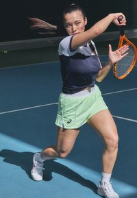 Tennisspelare i en marinblå och vit topp, ljusgröna shorts och vita skor, som svingar ett racket på en blå bana.