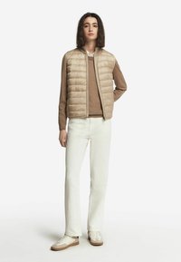 Beige pufferväst med quiltat mönster, brun tröja under, vita byxor och ljusa sneakers. Enkel, modern design.