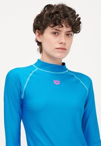 Rash guard azul de mangas longas feito de tecido elástico. Apresenta um logótipo rosa e costura em contraste. Gola alta e design justo.