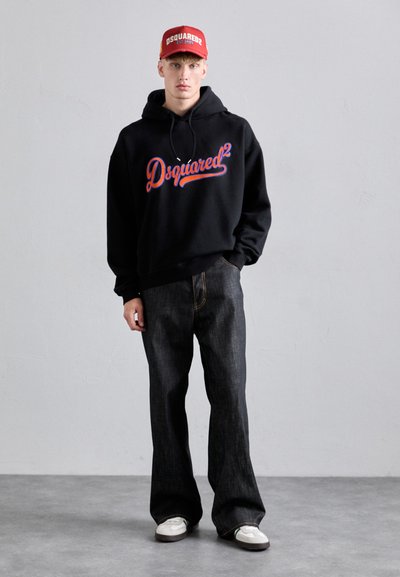 Schwarzer Hoodie mit rotem und blauen "Dsquared2" Logo, kombiniert mit dunklen Jeans. Rote Kappe mit Logo. Weiße Sneakers setzen einen Kontrast.