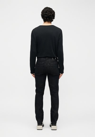 Άνδρας με σγουρά μαλλιά που φοράει μαύρο μακρυμάνικο πουκάμισο, μαύρα τζιν με σχέδια και παπούτσια πλατφόρμα, στέκεται με την πλάτη προς το μέρος μας σε απλό φόντο.