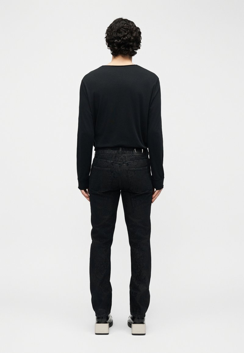 Uomo con capelli ricci che indossa una maglia nera a maniche lunghe, jeans neri con motivo e scarpe con plateau, in piedi di spalle su uno sfondo neutro.