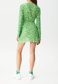 Femme debout, portant une robe mini à manches longues verte avec un motif floral blanc et des baskets à plateforme blanches avec des chaussettes teal.