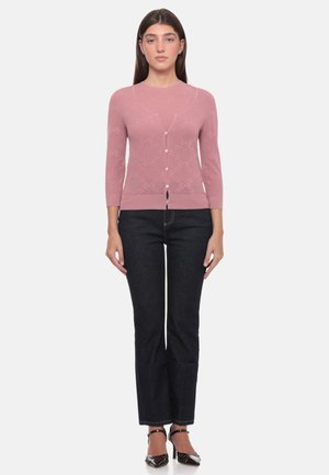 Donna in piedi rivolta in avanti che indossa un cardigan rosa chiaro con bottoni, jeans scuri e tacchi neri con cinturino alla caviglia su sfondo bianco.