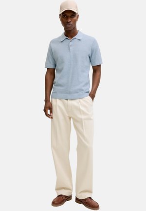 Mężczyzna w jasnoniebieskiej polo z krótkim rękawem, kremowych spodniach o szerokich nogawkach, brązowych butach i beżowej czapce z daszkiem, stojący z jedną ręką w kieszeni.