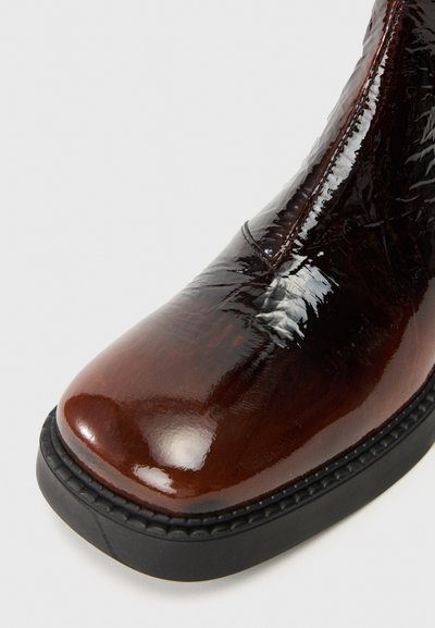 Καφέ λουστρίνι μπότες μέχρι τον αστράγαλο με στρογγυλή μύτη, γυαλιστερή επιφάνεια και μαύρη ελαστική σόλα με ραμμένες λεπτομέρειες κατά μήκος της άκρης.