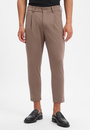 Man draagt cropped taupekleurige geplooide broek met zwarte leren loafers en een zwart overhemd tegen een witte achtergrond.