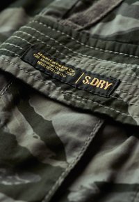 Camouflage stof in tinten groen en grijs, met zichtbare stiksels. Heeft een label met de tekst: "Manufacturing Co / S.DRY" in goud op een zwarte achtergrond.