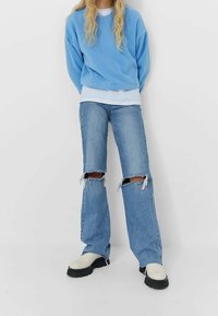 Felpa oversize azzurro chiaro, indossata sopra una t-shirt bianca, abbinata a jeans azzurro chiaro strappati con ampie aperture sulle ginocchia e stivali color crema.