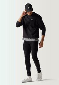 Sudadera negra con logo blanco, camiseta gris en capas debajo, pantalones ajustados negros y zapatillas blancas, rematada con una gorra negra con logo.