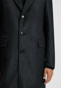 Cappotto grigio scuro con motivo pied de poule, caratterizzato da un colletto a punta, tre bottoni, due tasche laterali e tessuto testurizzato.