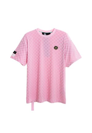 Mujer rubia con una camiseta rosa de cuadros suelta y de gran tamaño y pantalones anchos a juego, de pie con una mano en el bolsillo sobre fondo blanco.