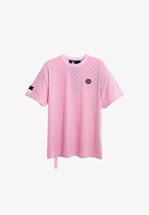 Mujer rubia con una camiseta rosa de cuadros suelta y de gran tamaño y pantalones anchos a juego, de pie con una mano en el bolsillo sobre fondo blanco.