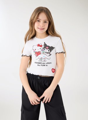 Ragazza giovane con i capelli lunghi sorridente, che indossa una maglietta bianca con i personaggi di Hello Kitty e la scritta "GLI AMICI sono dove c'è il DIVERTIMENTO", e pantaloni neri.