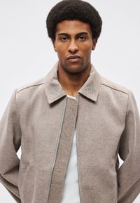 Beżowa kurtka z mieszanki wełny z zapięciem na zamek i złożonym kołnierzem. Posiada haftowane akcenty na kołnierzu oraz gładką teksturę.