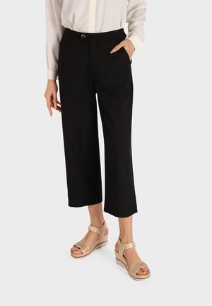 Donna con pantaloni neri a gamba larga e corti, camicetta bianca a maniche lunghe e sandali beige con zeppa e cinturini alla caviglia.