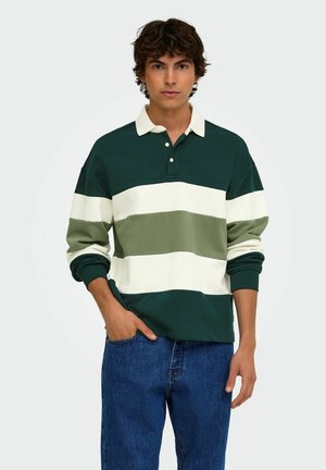 Giovane uomo con capelli castani ricci indossa una maglia da rugby a righe verdi, bianche e verde chiaro e jeans blu, in piedi con una mano in tasca.