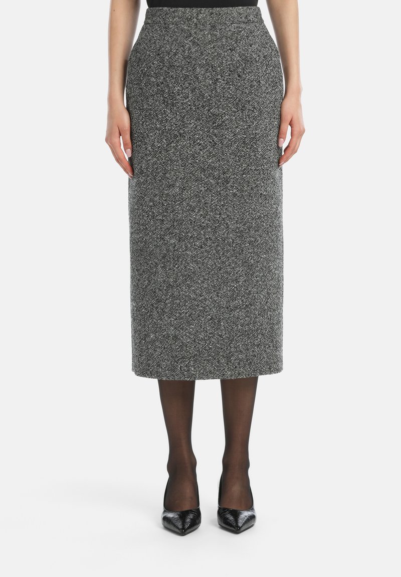Egy nő magas derekú szürke tweed midi szoknyát, fekete átlátszó harisnyát és fekete hegyes orrú magassarkút visel, egy fehér háttér előtt �áll.