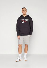 Svart Reebok-hoodie med en stor logotyp, kombinerad med grå shorts. Tyget verkar slätt, och outfiten kompletteras med vita sneakers.