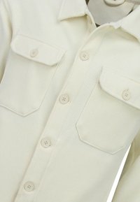 Lichtbeige overhemd met knopen, ribbelige structuur, twee borstzakken en standaardkraag. Voorzien van bijpassende knopen aan de voorkant.