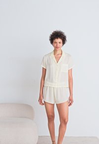 Vaaleankeltainen lyhythihainen pyjama-setti, jossa on napitettavat yläosa ja yhteensopivat shortsit, valmistettu pehmeästä ja kevyestä kankaasta. Sisältää rintataskun.