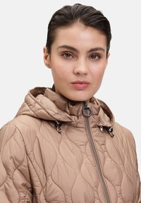 Jasno beżowa pikowana kurtka z kapturem, z metalowym suwakiem i akcentami w postaci sznureczków. Materiał wydaje się miękki i gładki z falistym wzorem.