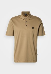 Béžové polo tričko z mäkkej bavlny; má zapínanie na tri gombíky a malé čierne logo na ľavej strane hrudníka; krátke rukávy.