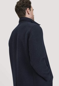 Man met kort bruin haar, gekleed in een donkerblauwe jas met hoge kraag, van achteren afgebeeld terwijl hij naar rechts kijkt.