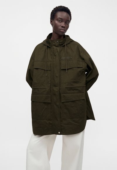 Personne grande aux cheveux courts portant une veste à capuche vert foncé oversize avec de grandes poches et un pantalon blanc à jambes larges.