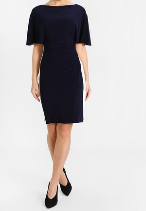 Kvinde iført en knælængde marineblå kjole med hvide sidepaneler og sorte spidse hæle, stående med korslagte ben mod en hvid baggrund.